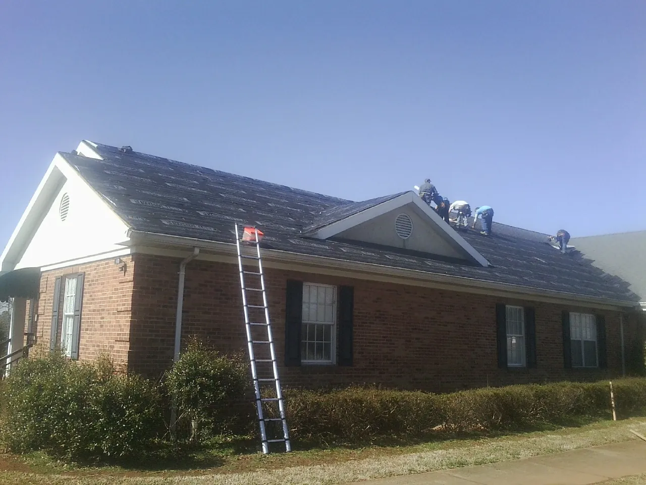 Crew tearing shingles off a brick ranch roof with a ladder set — Metal Roof Repair work by our crew in Dimondale