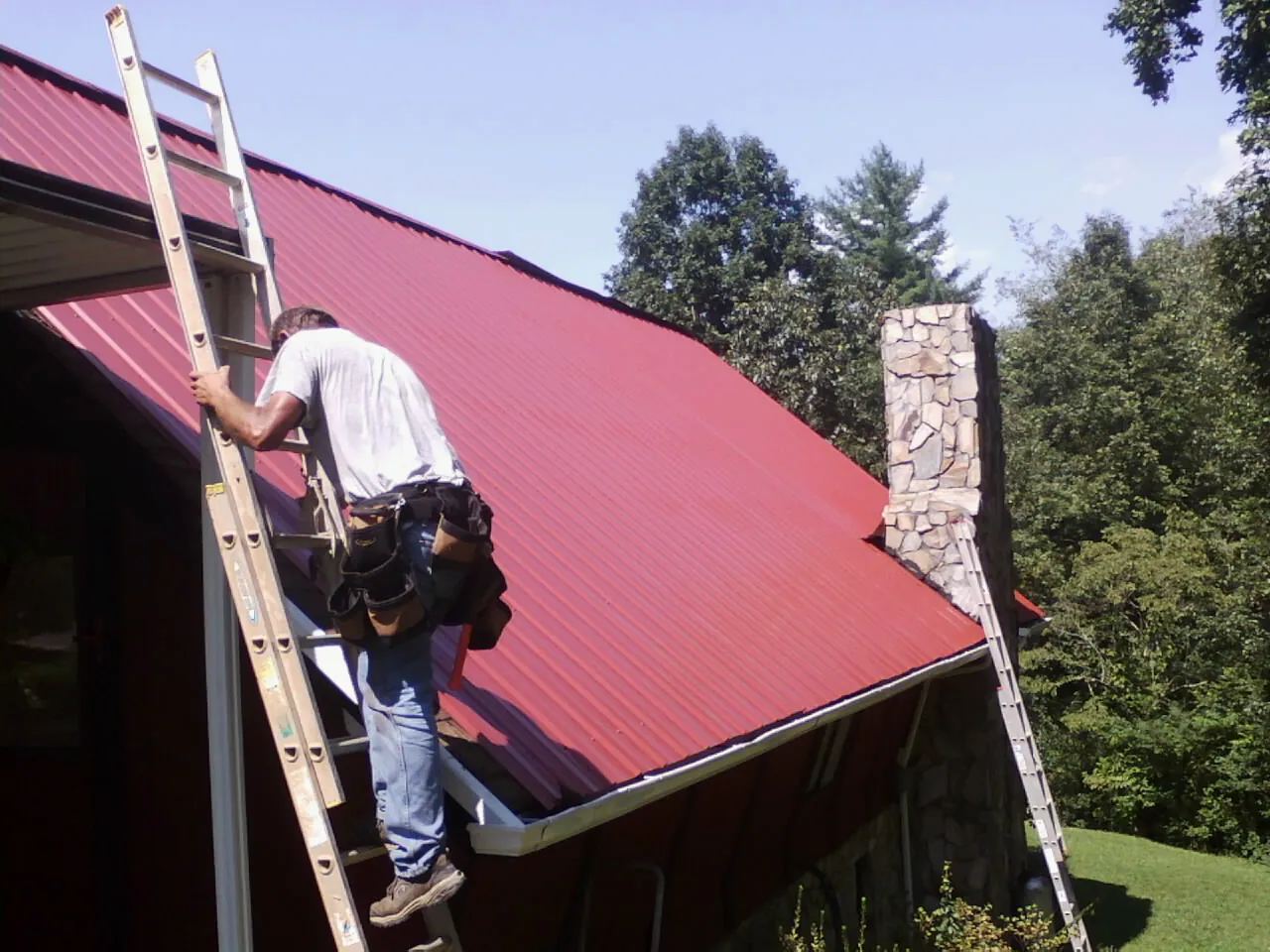 Roofer on a ladder beside a red metal roof and stone chimney — Soffit & Fascia Repair by Roofing Brothers in Dimondale